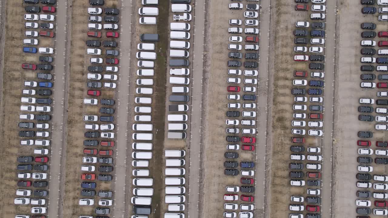 An aerial video of a large parking lot featuring neatly arranged rows of parked cars. The vehicles are aligned in long sections, showcasing a variety of colors and types on a spacious outdoor lot.