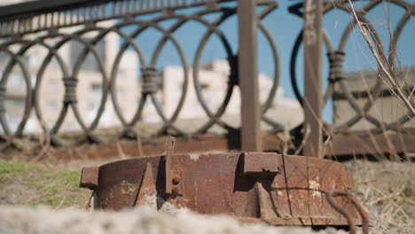 close view of a rusty metal object lying on the ground with an ornate urban fence and a blurred white car in motion with other cars