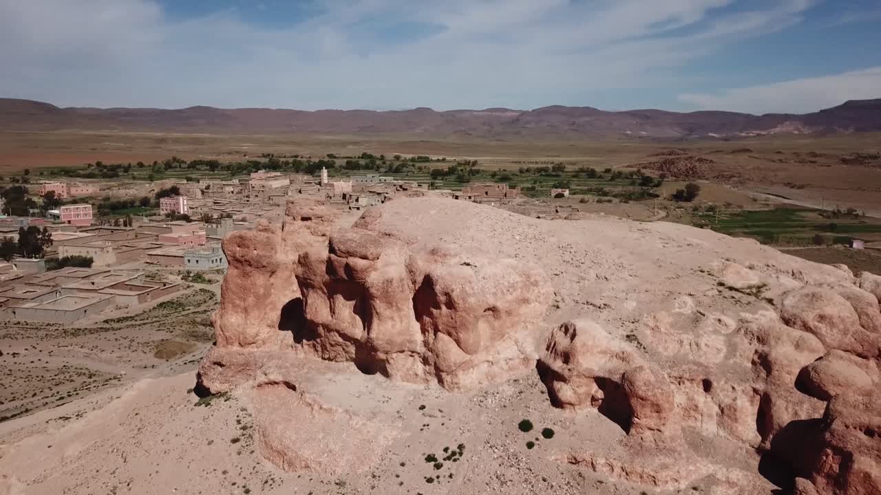 Aerial drone shot of a unique red rock formation shaped by erosion in Ouarzazate. The geological landmark stands near a village in the arid Moroccan desert landscape