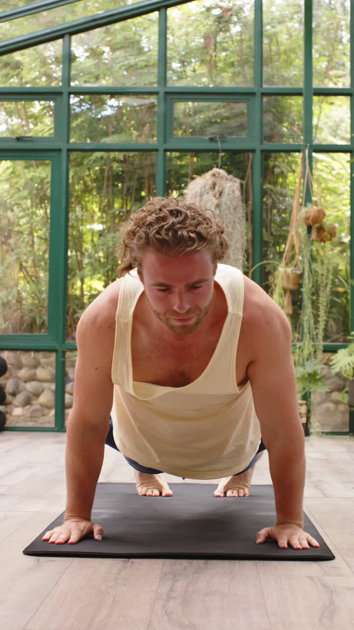 Vertical video: Practicing yoga, man in upward dog pose on mat in glass house