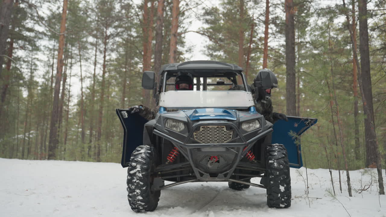 Controllo pre-corsa di un ATV nella foresta innevata. Due piloti con casco e in mimetica aprono gli sportelli e ispezionano un robusto buggy side-by-side, controllando pneumatici, paraurti e parabrezza prima della partenza, in un paesaggio di pini freddo e nitido.