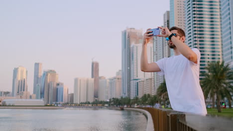 Ein-Junger-Männlicher-Tourist-Mit-Sonnenbrille-Und-Borstigem-Hipster-In-Einem-Weißen-T-Shirt-Steht-Am-Wasser-Vor-Dem-Hintergrund-Der-Modernen-Stadt-Und-Macht-Fotos-Und-Videos-Auf-Dem-Telefon-Für-Soziale-Netzwerke,-Die-Live-übertragen-Werden