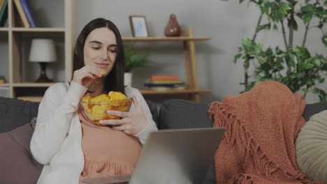 pregnant woman snacking on chips and watching laptop