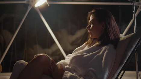 pensive woman in bathrobe reclines in armchair spending time on home terrace. sombre lady thinks important issues over relaxing in room with subdued lighting