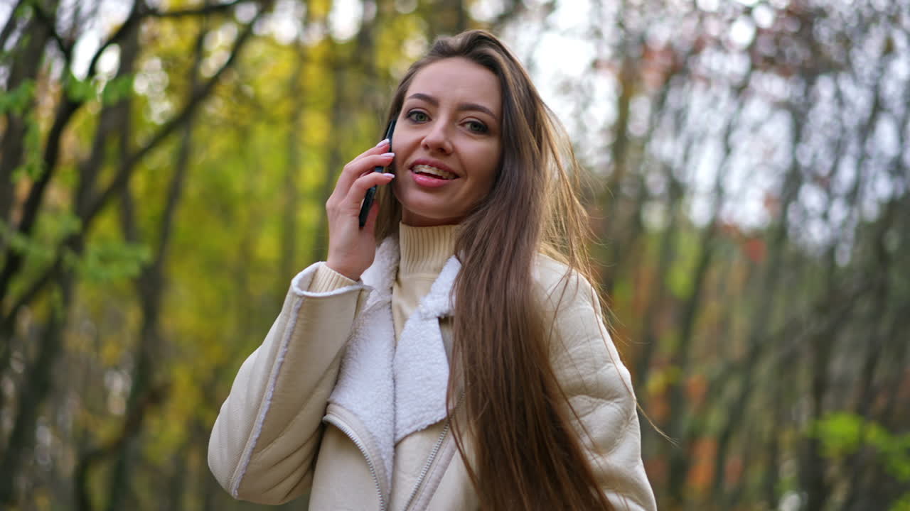 Positive attractive brunette speaking on the phone smiling outdoors. Lady touches her hair looking into camera and holding her phone at the ear.