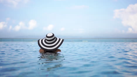 a woman with her back to the camera wearing a floppy black and white sunhat is neck-deep in a resort swimming pool looking out to the ocean