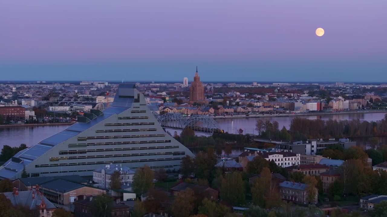 Drone sorvola Riga al tramonto, mostrando la luna piena e un treno su un ponte