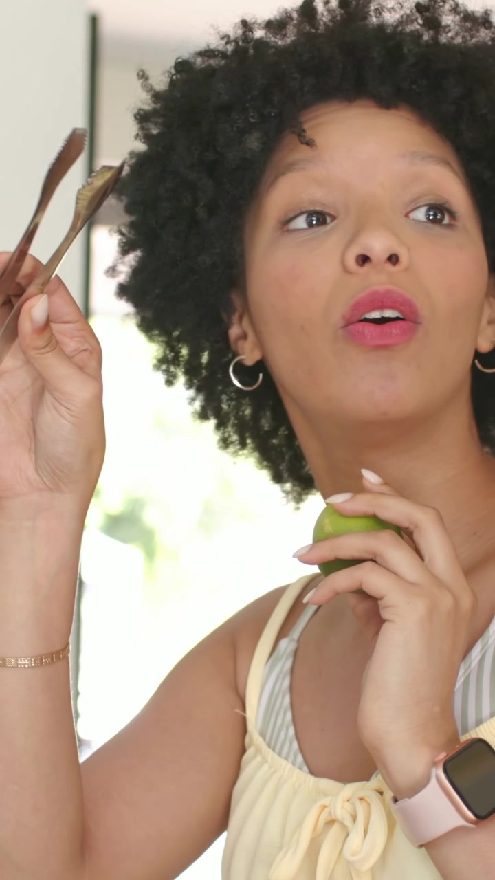 Vertical video: Smiling woman replying to offscreen cue, holding apple, tongs, gesturing in kitchen