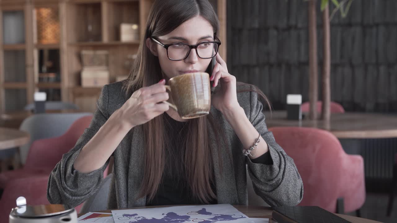 bella joven empresaria con anteojos está sentada en un café, tomando un café, hablando por teléfono y estudiando algunos diagramas y estadísticas