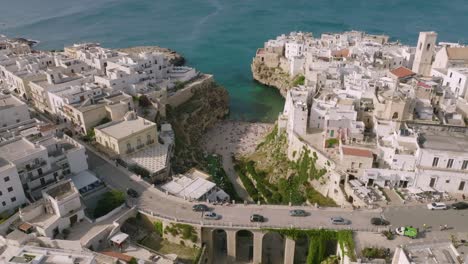 Sobrevuelo-Aéreo-Lento-Del-Tráfico-De-Automóviles-Que-Pasa-Por-Un-Puente-En-Polignano-A-Mare,-Italia-Con-Vistas-Al-Mar-Y-La-Playa