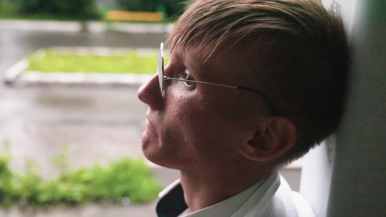 Biologist in white coat leans head against wall during calm urban break, chewing pastry with thoughtful expression while enjoying quiet outdoor moment surrounded by greenery and wet pavement