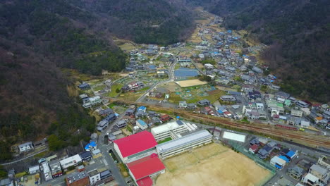 Aerial-View-of-Hinase-City,-Okayama,-Japan:-Stunning-Top-Down-Shot-of-School-and-Field,-Pan-Out-to-Show-Charming-City-with-Small-Buildings,-Green-Mountains,-White-Sky