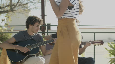 two men sitting and playing guitar during a rooftop party while an unrecognizable woman holding beer and dancing