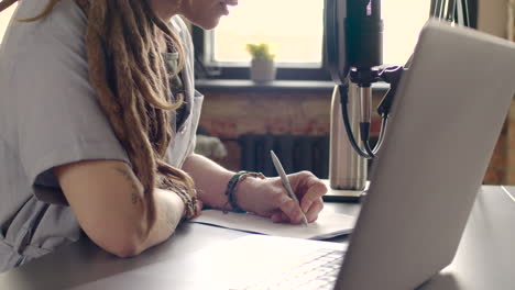 le mani della donna prendono appunti durante la registrazione di un podcast seduto su un tavolo con laptop e microfono