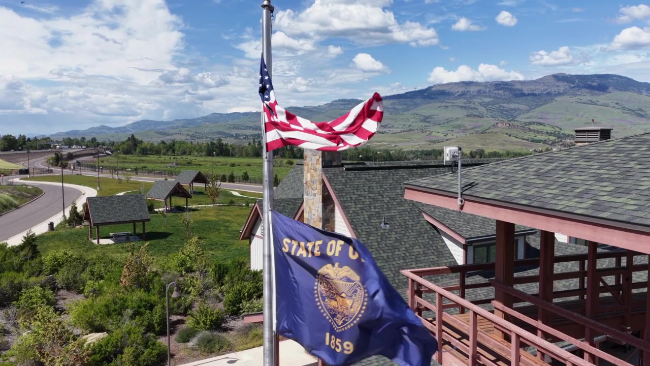 American flag flies beside state flag atop welcome center rooftop in rural Oregon, USA, overlooking green valley, winding road, distant mountains under partly cloudy sky. Aerial drone view.