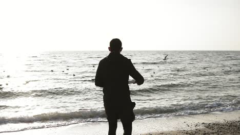 Young-sportive-male-with-caucasian-appearance-in-black-sports-outwear-running-along-the-seashore-with-headphones-plugged-in.-Beautiful-scenic-view