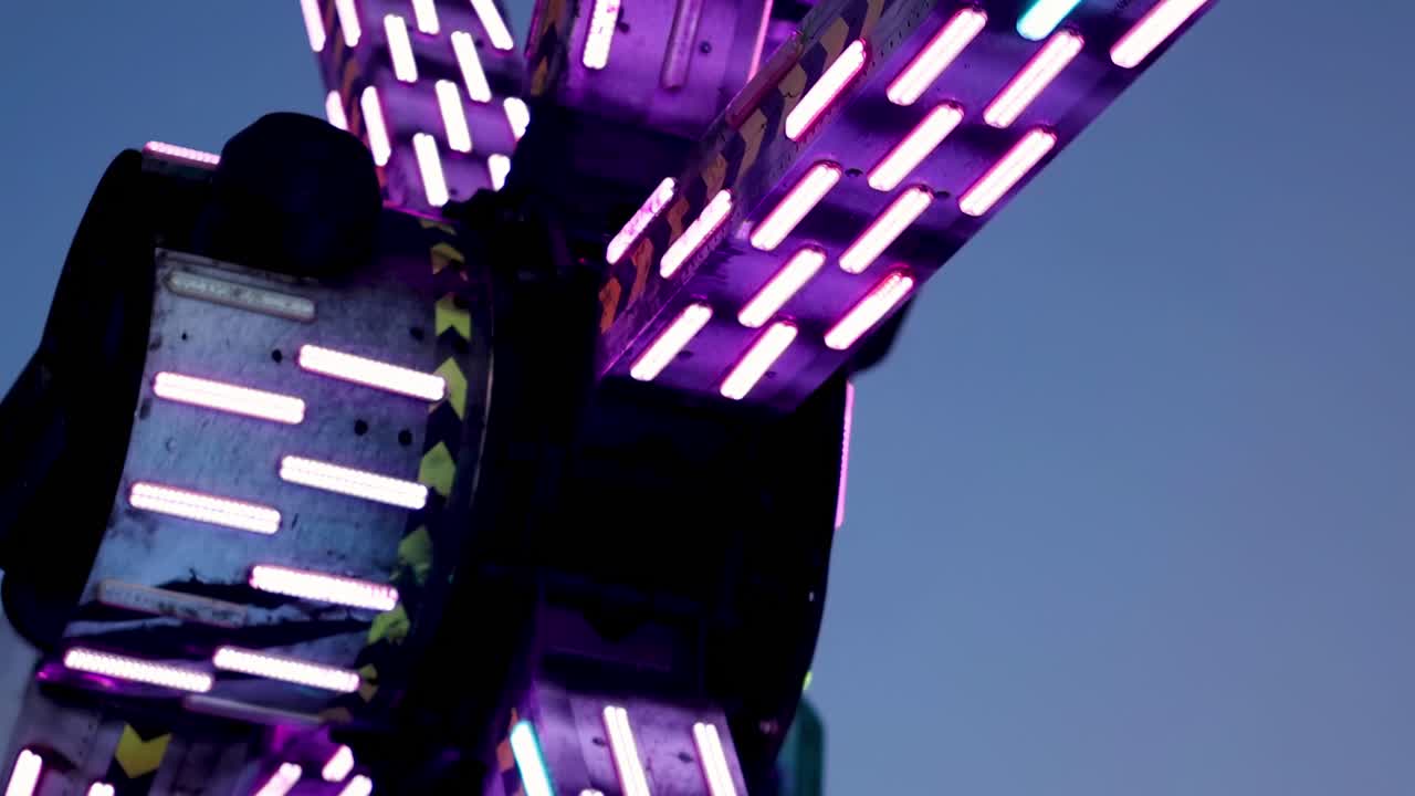 Close-up of illuminated mechanical arms with neon lights against a clear sky, showcasing vibrant colors and intricate design.
