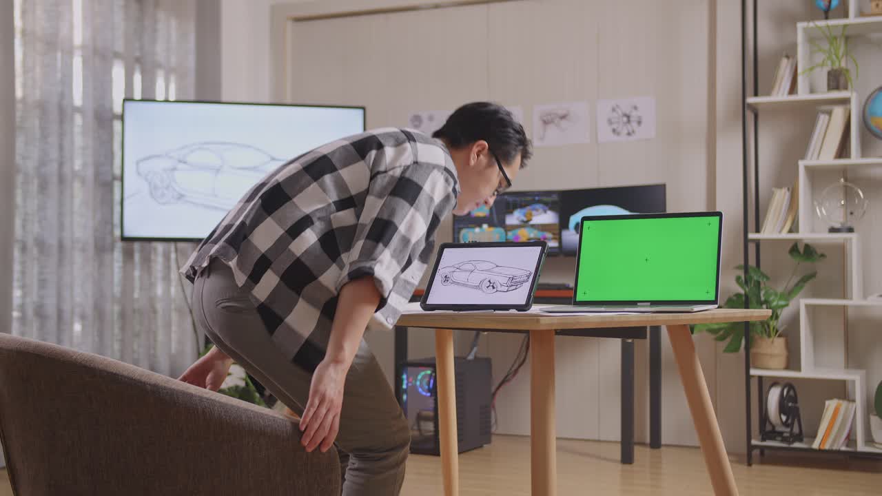 아시아 남성이 앉아서 타블에 자동차 컨셉을 그려내고 있는 측면 뷰 tv와 컴퓨터를 가진 스튜디오에서 녹색 스크린 노트북 3d 전기 자동차 모델을 표시합니다.