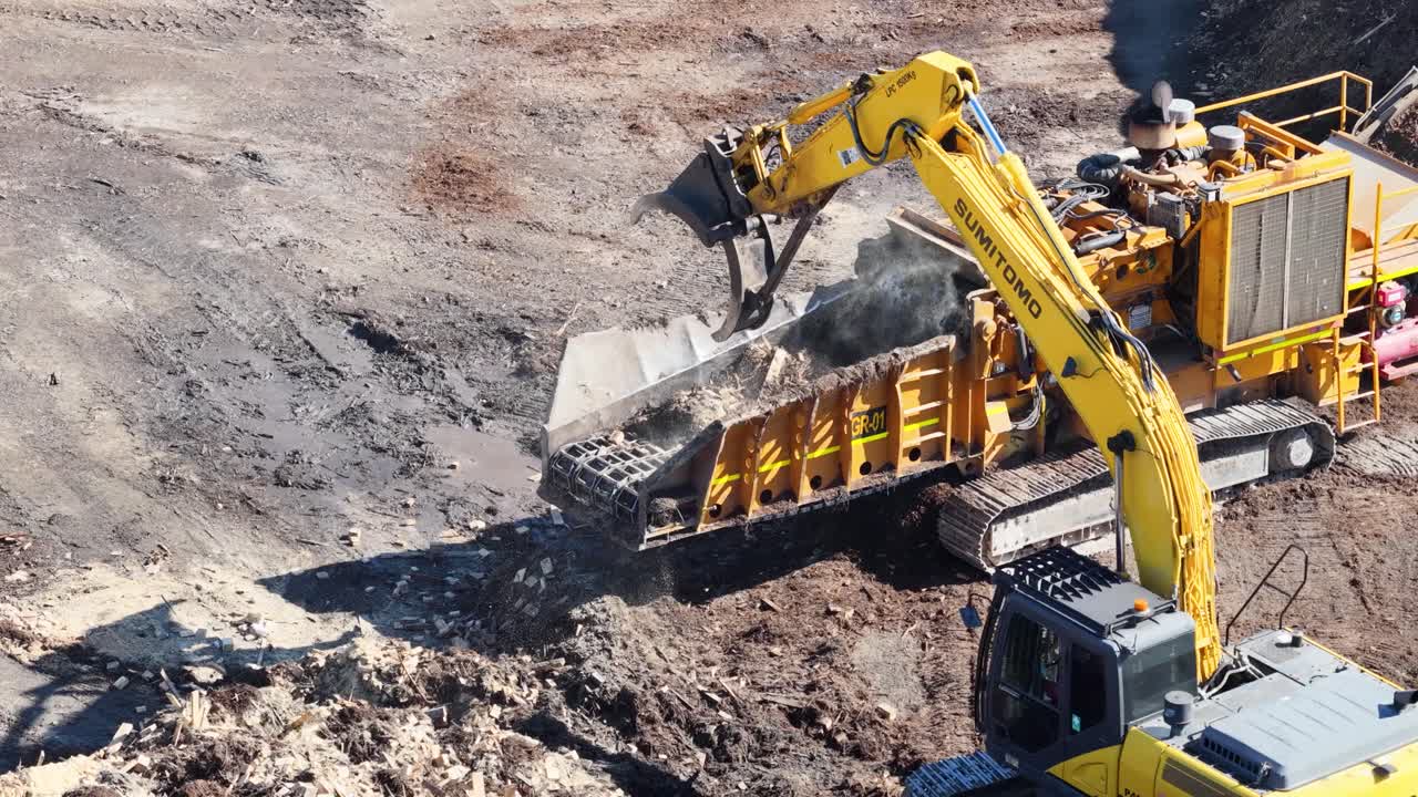 Drone captures an excavator transferring soil into a processing machine in a sunlit, industrial environment