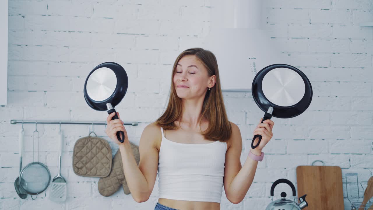 Young female with frypan in the kitchen. Attractive housewife holding two new nonstick pans and closing her face by them. Happy woman in the modern kitchen.