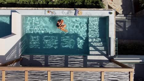 Lovely-couple-enjoying-the-pool-at-Cayo-Resort-in-Greece,-Crete---man-holds-and-turns-brunette-woman-in-swimming-costume-in-water---drone-flies-in-bird's-eye-view-over-them