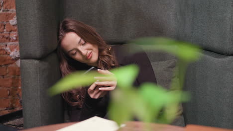 young woman in black outfit holds smartphone while leaning her head against green cushion. seated comfortably with relaxed posture in cozy indoor setting, soft plant blur in foreground adds depth