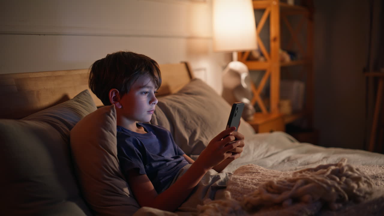 Child watching phone nighttime lying in cozy bed closeup. Little boy scrolling