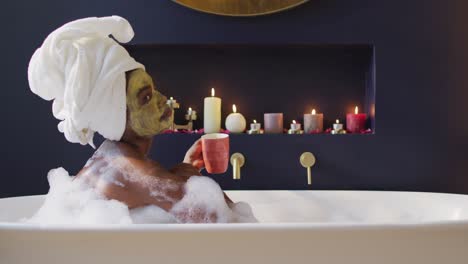 mujer afroamericana sonriente con toalla y máscara tomando un baño y bebiendo café en el baño