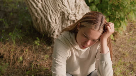 young lady seated on ground resting head on left hand, appearing lost in thought, with soft sunlight highlighting her expression, background includes textured tree bark and green foliage