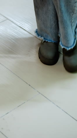 vista de la pierna de un individuo en vaqueros y botas llevando una bolsa de compras en la mano izquierda y una bolsa negra en la derecha, caminando en el centro comercial, sombra en el piso de azulejos