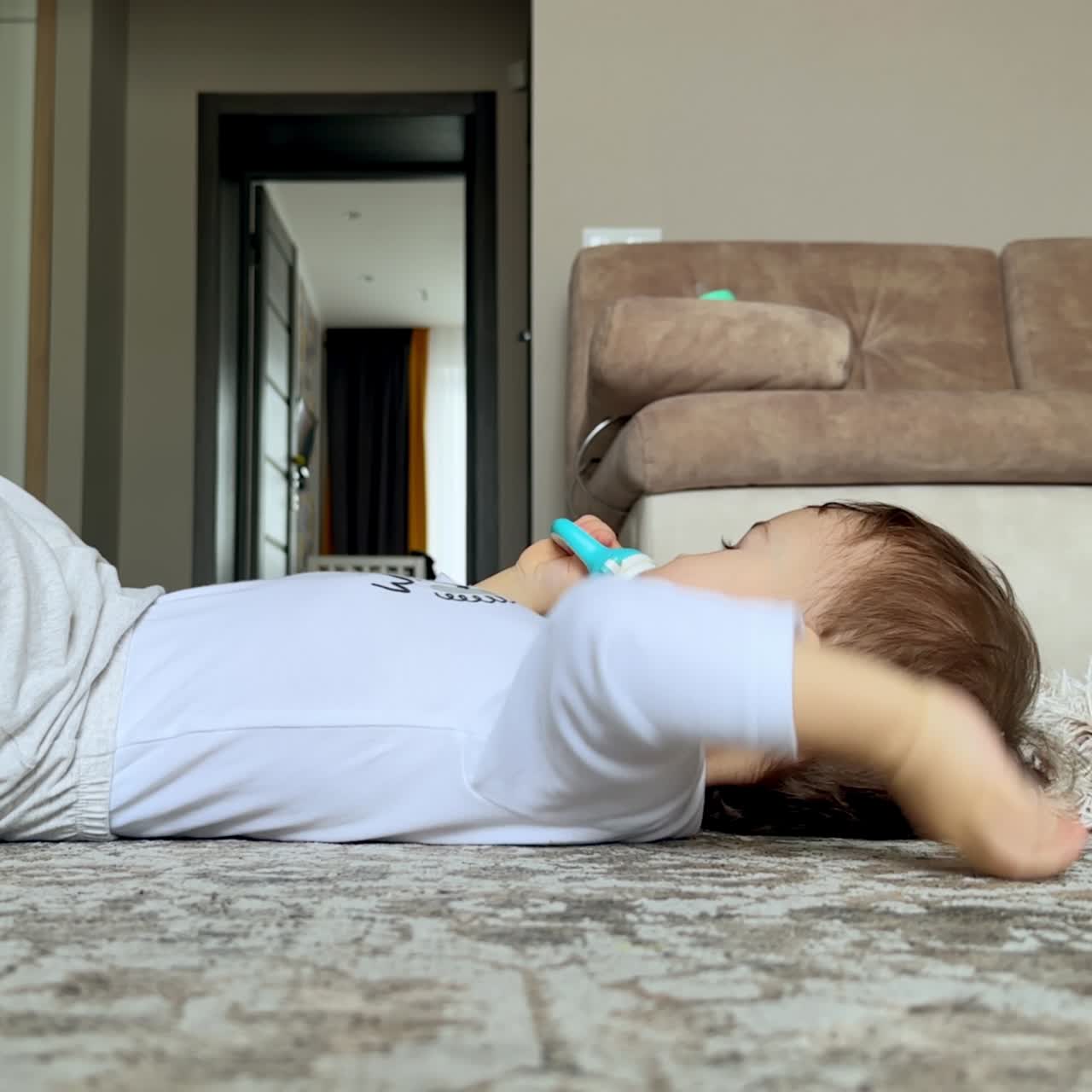 Child in white clothes lies on the floor holding a pacifier. Peaceful baby boy watches TV in the living room