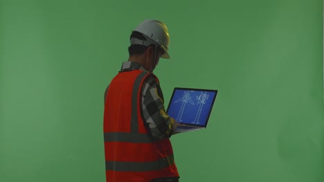 back view of asian male engineer with safety helmet looking at the wind turbine blueprint on laptop while standing in the green screen background studio, during sunset or sunrise time