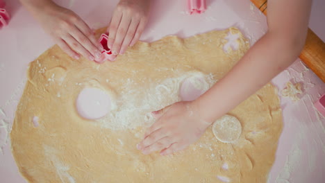 overhead view of kids pressing pink shape cutters into dough while one rubs flour across surface and quickly switches to cutting another shape, capturing playful baking moment on pink floured table