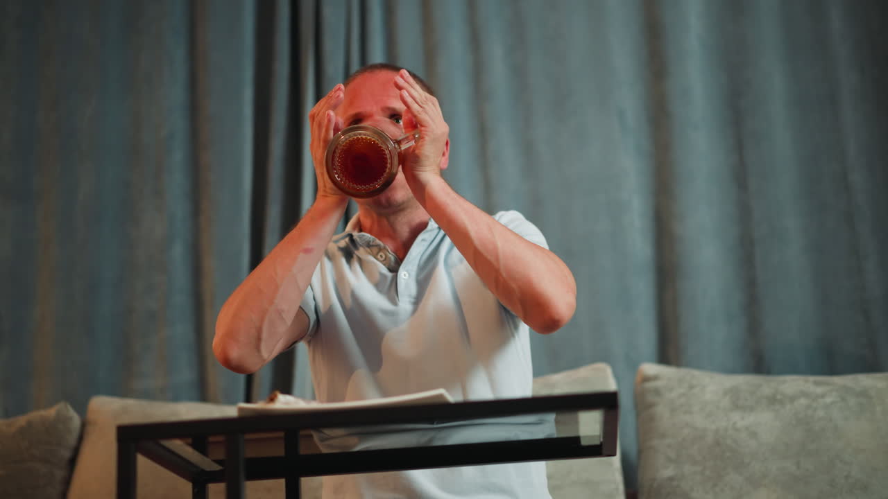 Young man seated at table lifts transparent mug filled with red berry drink, drinks thoughtfully, lowers mug with pensive expression, cozy background with curtains and pillows