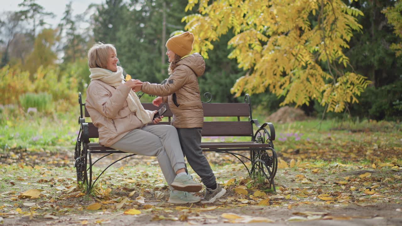 woman focusing on smartphone seated on park bench wearing pastel coat and knit scarf as child approaches holding bright yellow leaf then hugs woman warmly amid golden autumn foliage