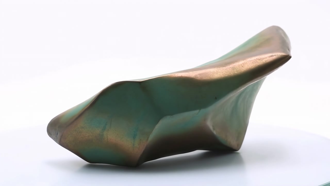 Reflective green metallic object gradually changes visible silhouette across frames while resting on white surface, showing smooth folds, curved edges, soft lighting, and minimalist presentation.