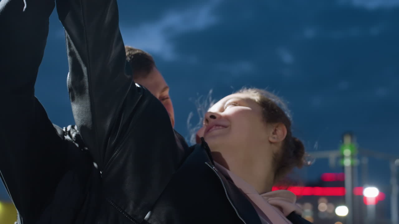 Close up of lovers dancing outdoors during windy evening with joyful expressions as hair blows gently in breeze and residential lights glow behind them under deep blue sky