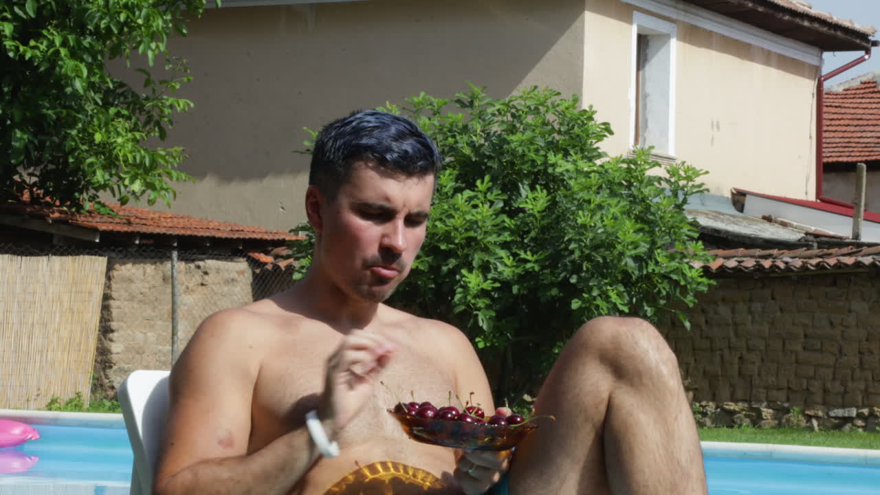 modelo masculino comiendo cerezas frescas mientras está sentado junto a una piscina al aire libre