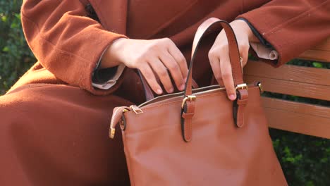 a woman wearing a brown coat sits on a bench and holds a brown leather handbag