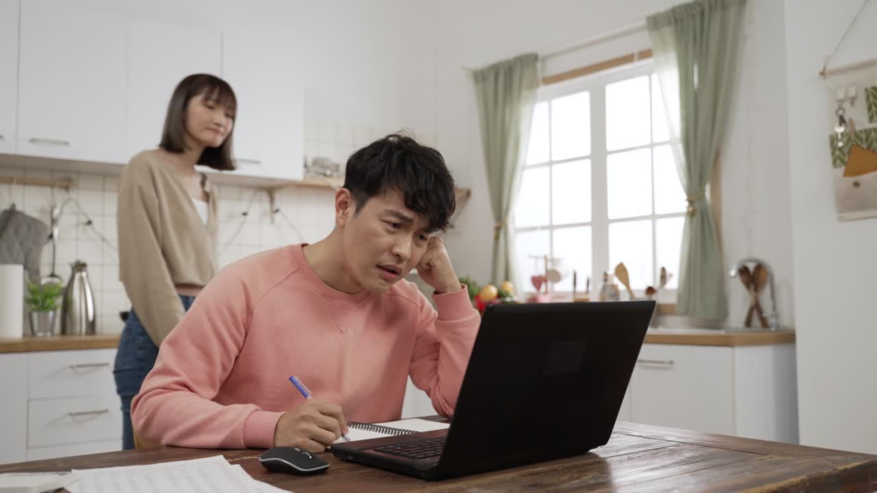 스트레스를 받은 아시아 남성이 노트북으로 청구서를 계산하고 소득세 신고를 온라인으로 하고, 아내가 다가와 그의 등에 박수를 치며 우려를 표한다.