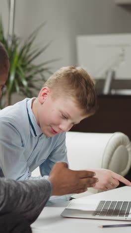 Little boy student listens and asks questions to African-American English teacher watching video lesson via laptop computer at home slow motion