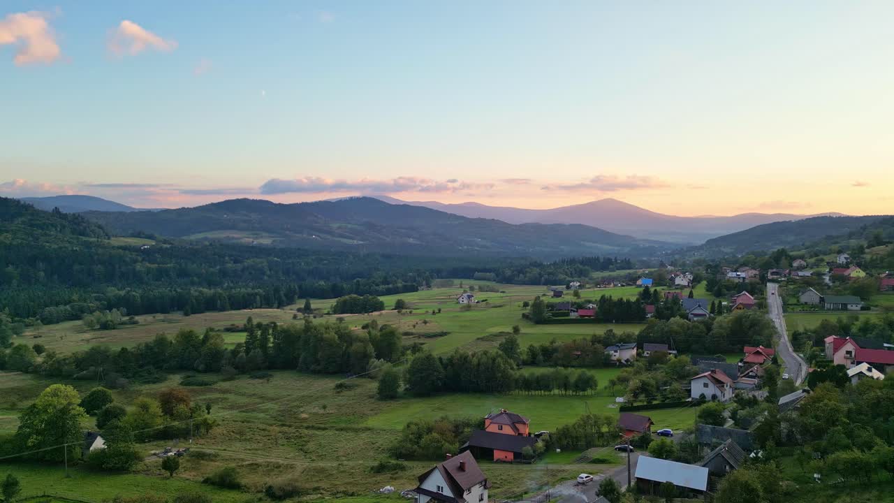 Aerial drone footage of a tranquil mountain village at sunset. Vibrant sky shifts from orange to yellow to blue, with scenic horizon and calm forward motion
