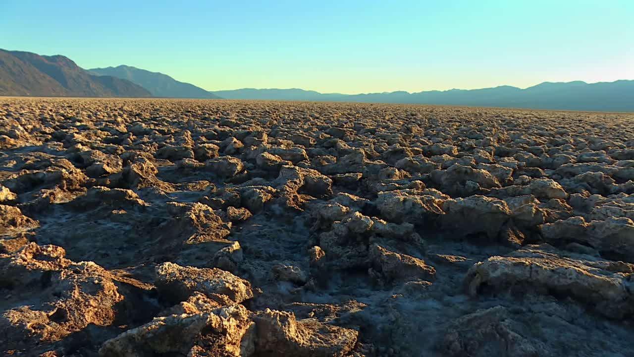 Wide view of jagged, crusty salt formations stretching across flat desert basin with distant mountains under golden light in Death Valley National Park, California, USA