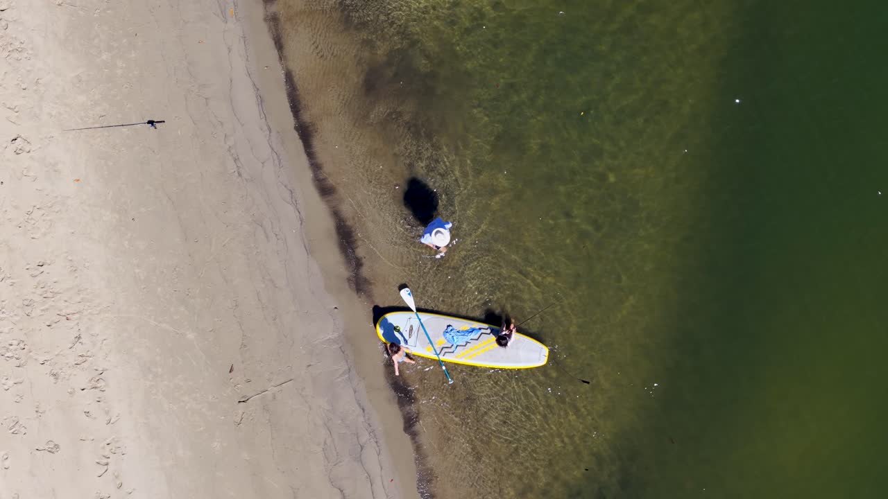 Two kids paddleboard and fish in shallow water, aerial view, bright daylight, relaxed atmosphere