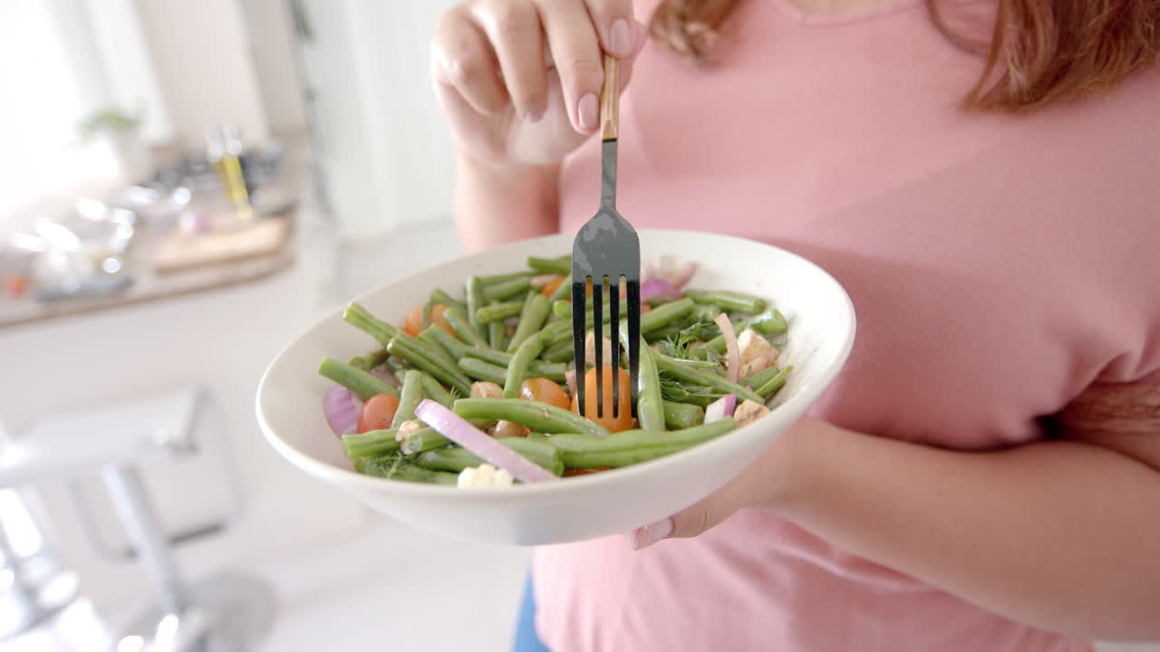 seção média de tamanho maior mulher biracial comendo salada de vegetais de pé na cozinha, em câmera lenta