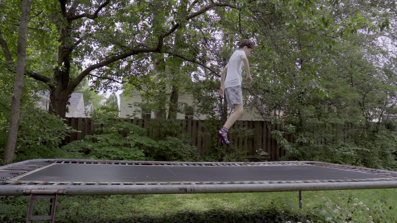 Cute boy jumps on a trampoline in the backyard, slow motion wide shot