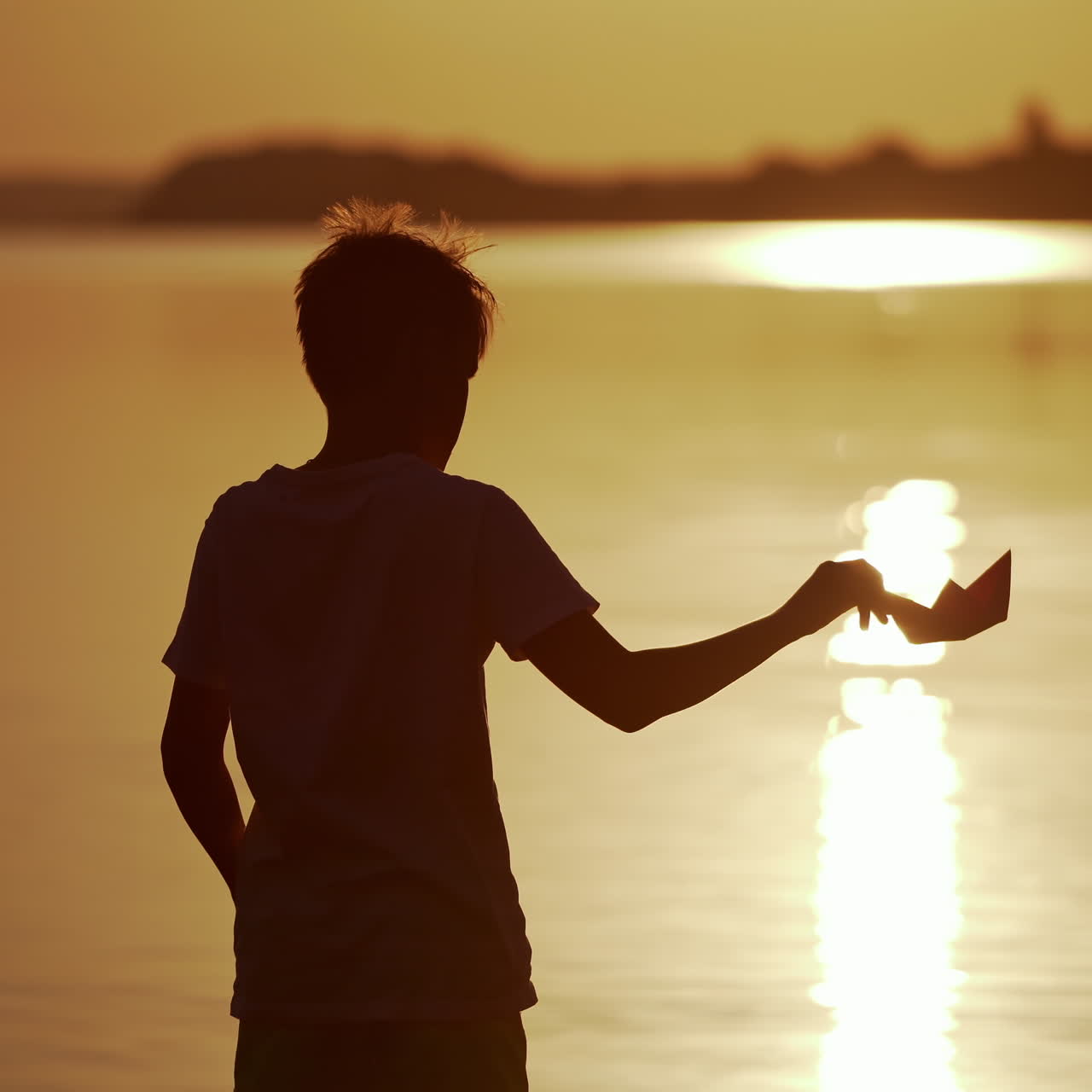 Silhouette of a boy with homemade origami ship in the evening. Child standing near the river at sunset and playing with paper boat in the air.