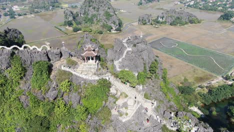 drone aerial view in vietnam flying down in front of rocky mountain covered with green trees with stairs and a temple in ninh binh on a sunny day