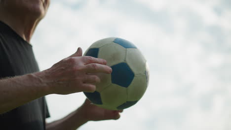 A side view of an elderly man gently rolling a soccer ball in his hand, with a cloudy sky seen
