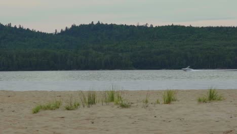 Speed-boat-and-pontoon-passing-by-camera-on-a-lake-beach-in-Lac-Taureau,-Quebec